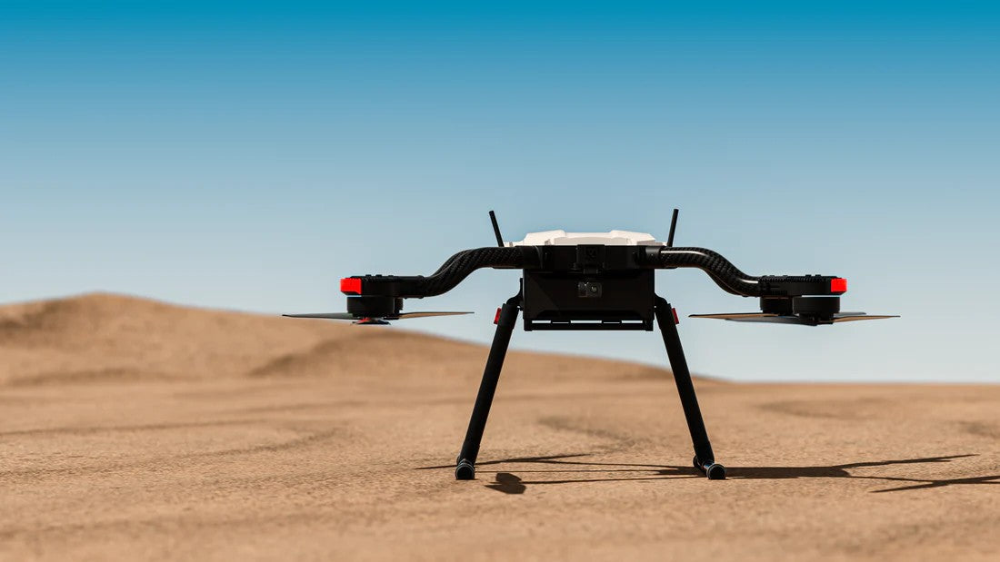 A multi-rotor drone sits atop a sunlit desert, with vast stretches of sand visible in the background.