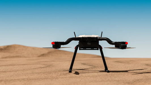 A multi-rotor drone sits atop a sunlit desert, with vast stretches of sand visible in the background.